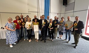 Gruppenfoto mit 6 Frauen und 10 Männern, die je eine Sonnenblume halten.