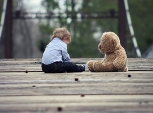 Symbolfoto: Kleinkind sitzt allein mit Teddybären auf einer Holzbrücke auf dem Spielplatz.