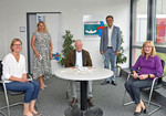 Foto: 5 Personen, davon 3 am Tisch sitzend und zwei stehend