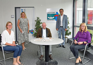 Foto: 5 Personen, davon 3 am Tisch sitzend und zwei stehend